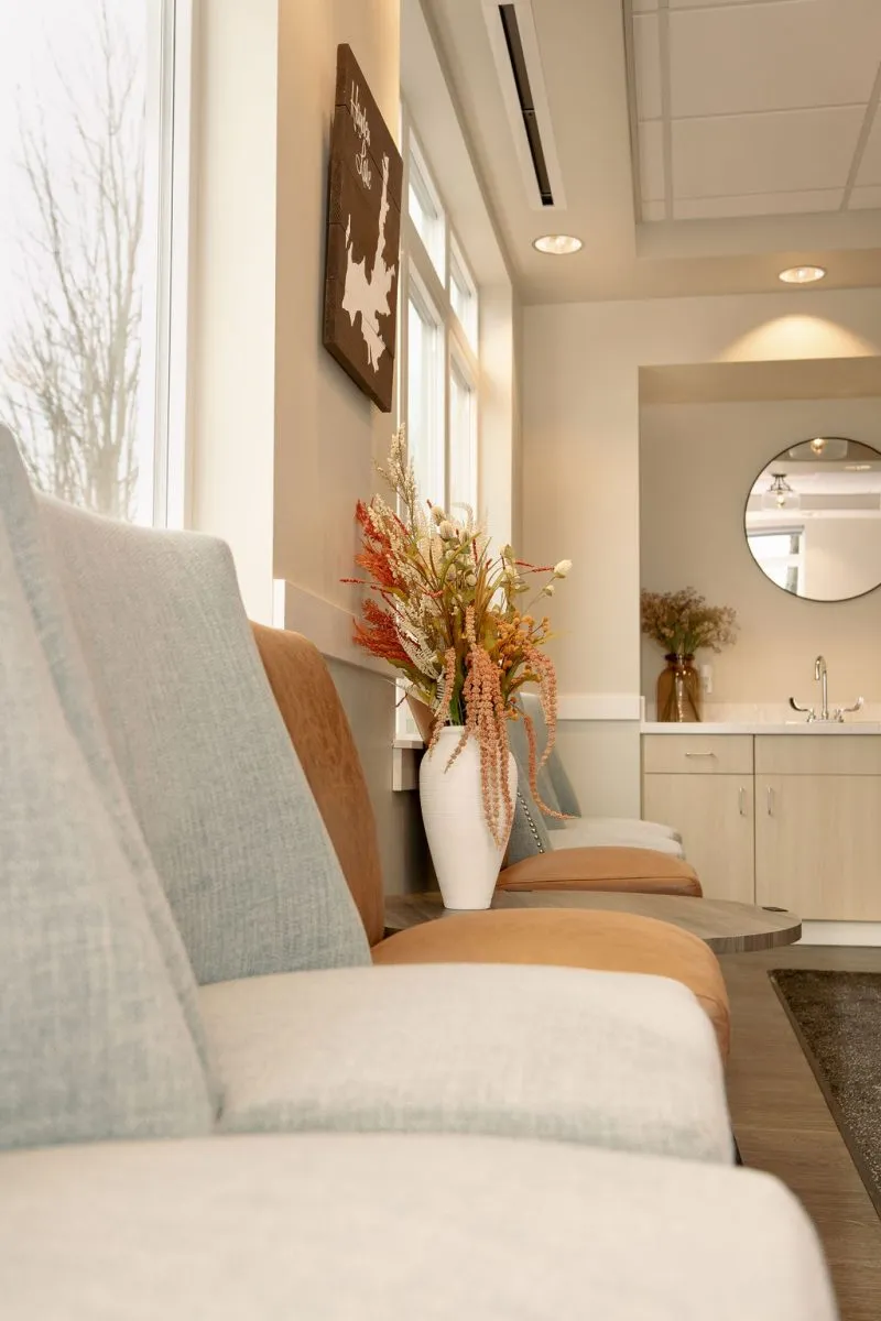 A cozy seating area in Dr. Trevor Griffitts' dental waiting room featuring neutral-colored cushions, a side table with a vase of dried flowers, and bright window lighting.