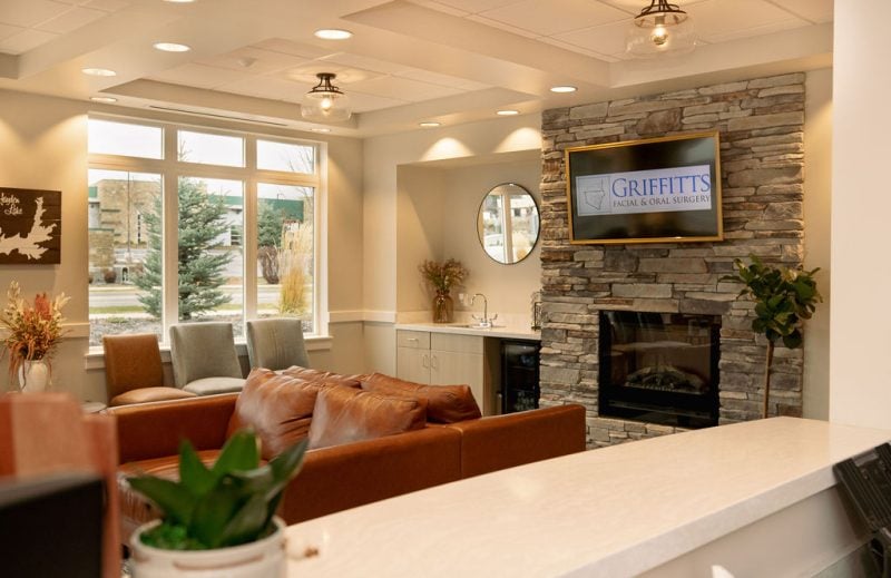 The comfortable reception area at Dr. Trevor Griffitts' dental office featuring a stone fireplace, wall-mounted television displaying the practice logo, and plush leather seating.