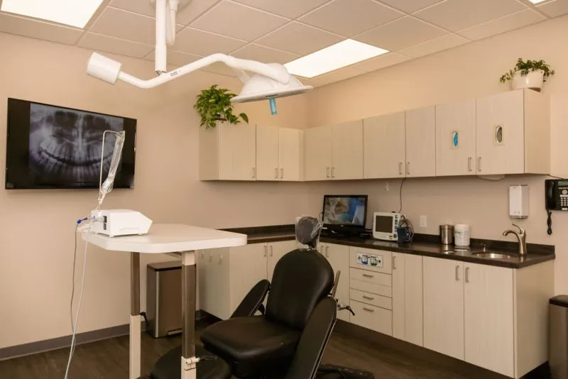 A modern dental examination room at Dr. Trevor Griffitts' practice equipped with a black treatment chair, overhead lighting, cabinets, and a digital X-ray screen on the wall.