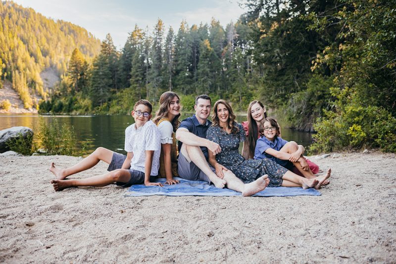 Dr. Trevor Griffitts, DMD, sitting outdoors on a blanket near a lake with his wife and four children for a family photo.